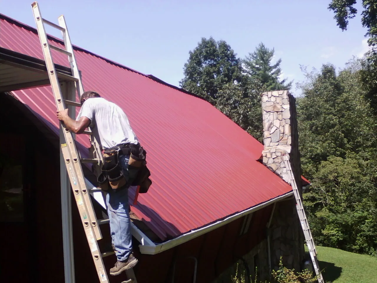Roofer on a ladder beside a red metal roof and stone chimney — Soffit & Fascia Repair by Roofing Brothers in Chugwater